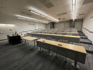 classroom seating showing location of stairs, shared table space, and accessible seating at front of room; a/v equipment is visible in the corner and on the ceiling