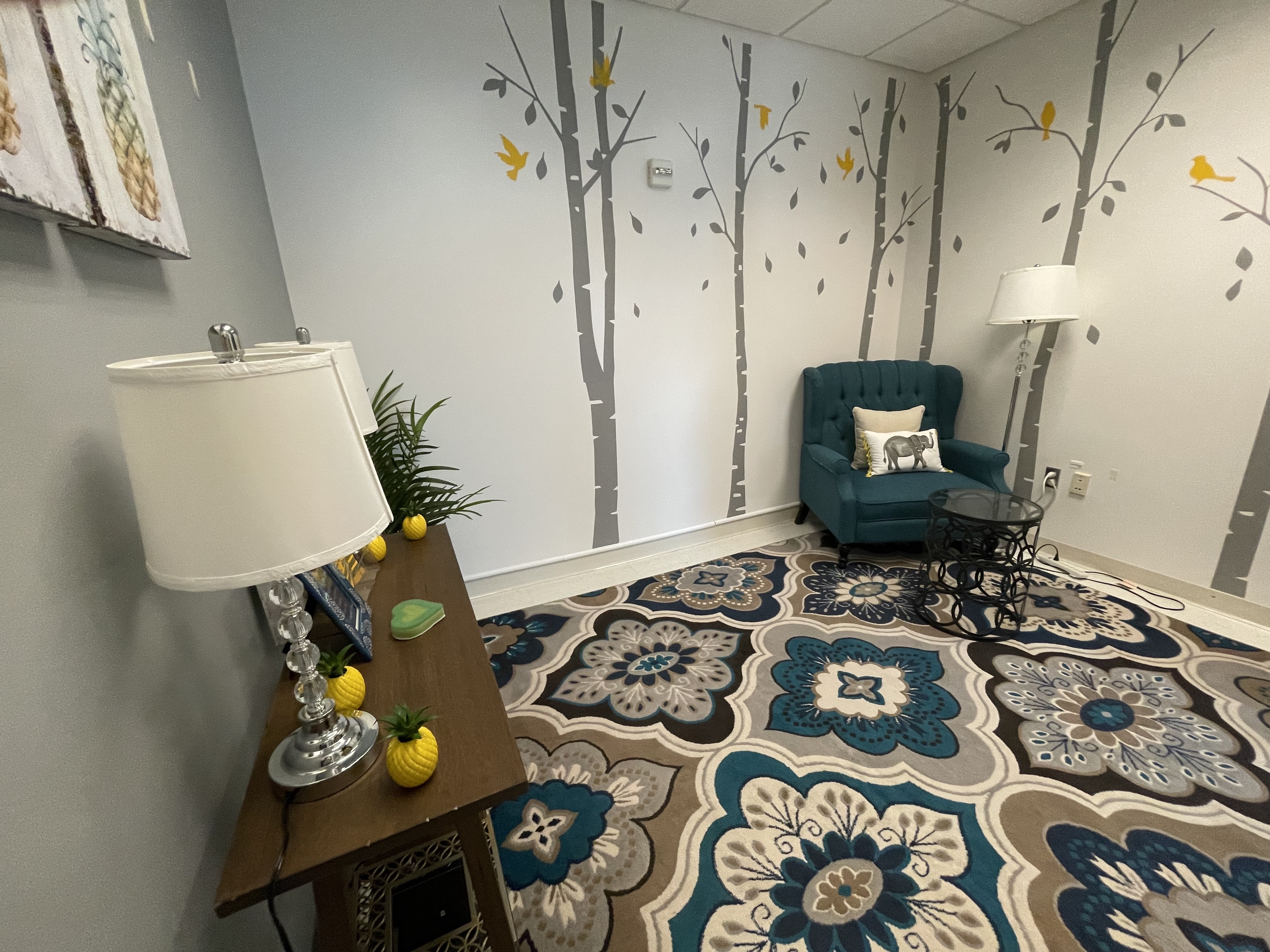 room with tree and bird murals on the wall, a colorful carpet, armchair, side table, and lamps